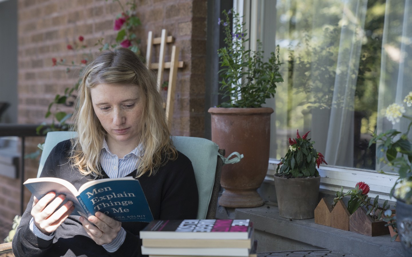 Student sitting out side on a patio reading with a stack of books on a table next to the student