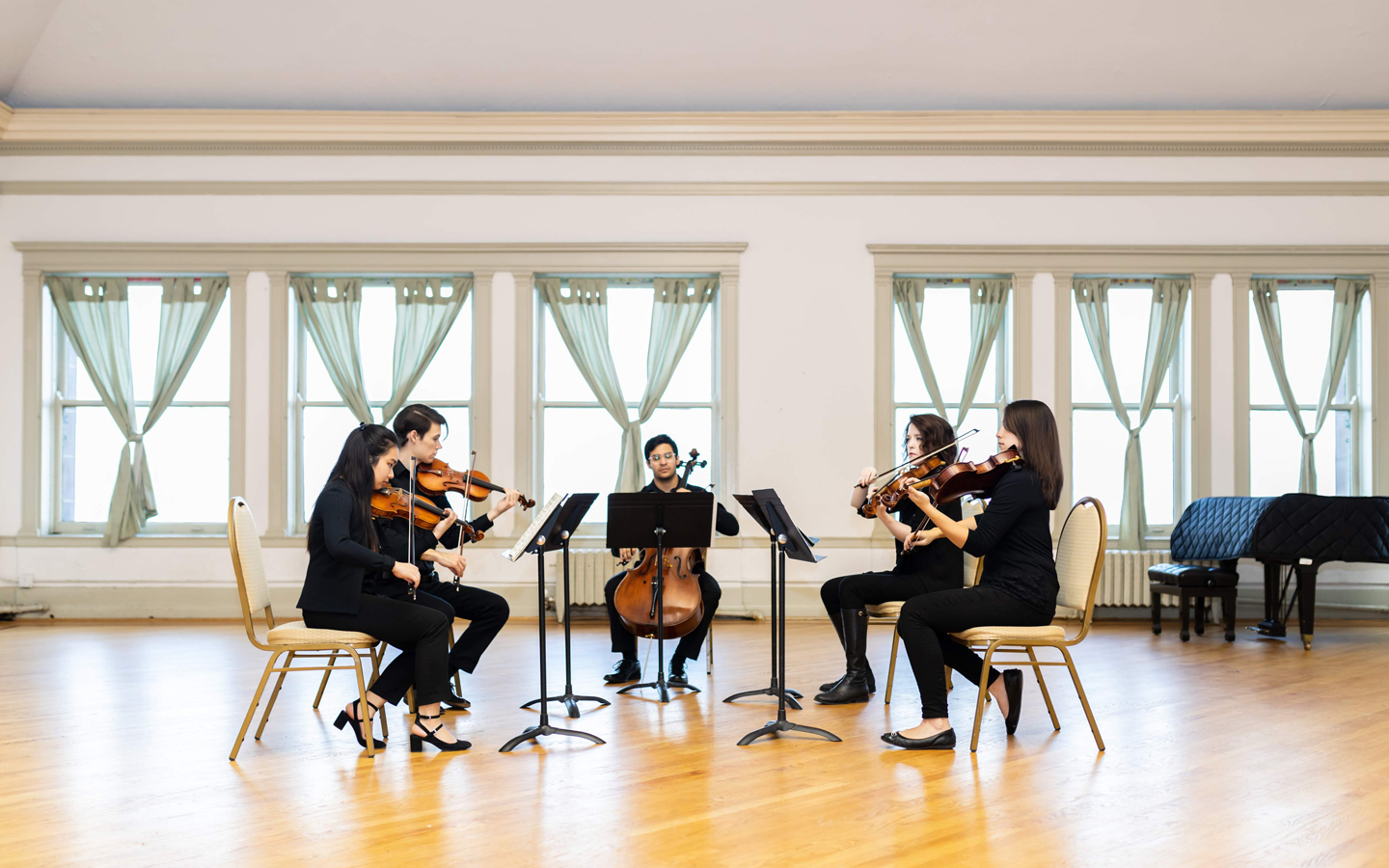 Four studentd playing violin and one student playing the cello sitting in a bright room with large windows.