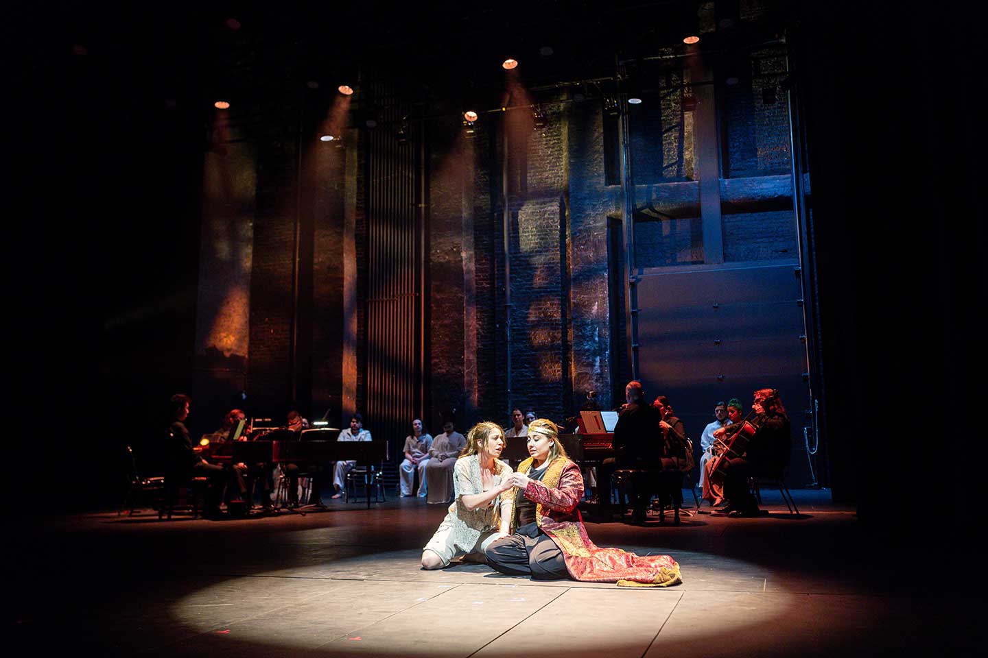 Long shot of two costumed performers kneel facing each other on a spotlighted stage, with musicians in the dimly lit background
