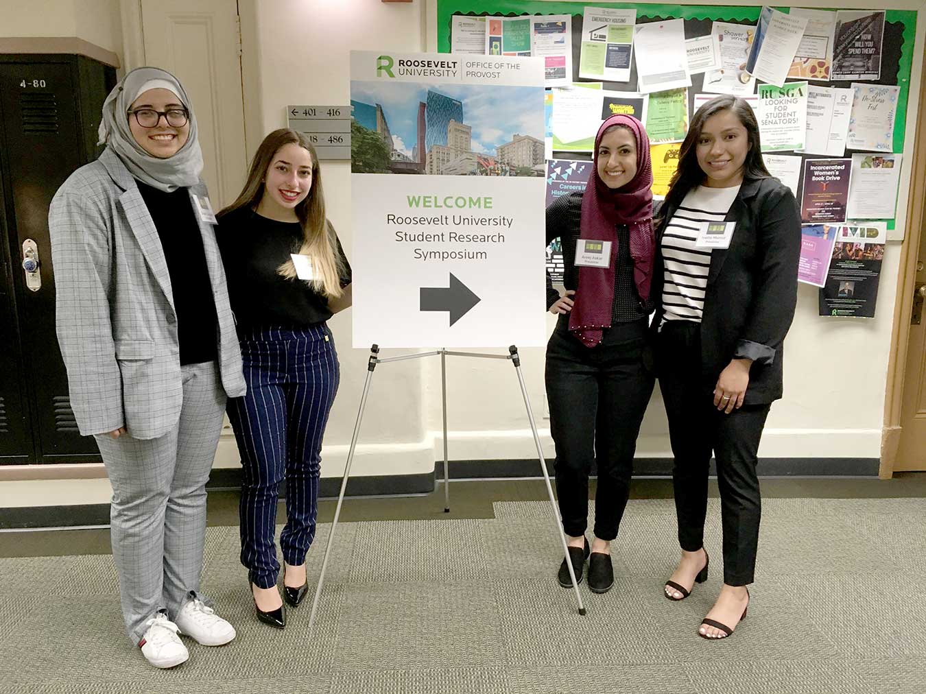 four students standing by a sign for the Student Research Symposium