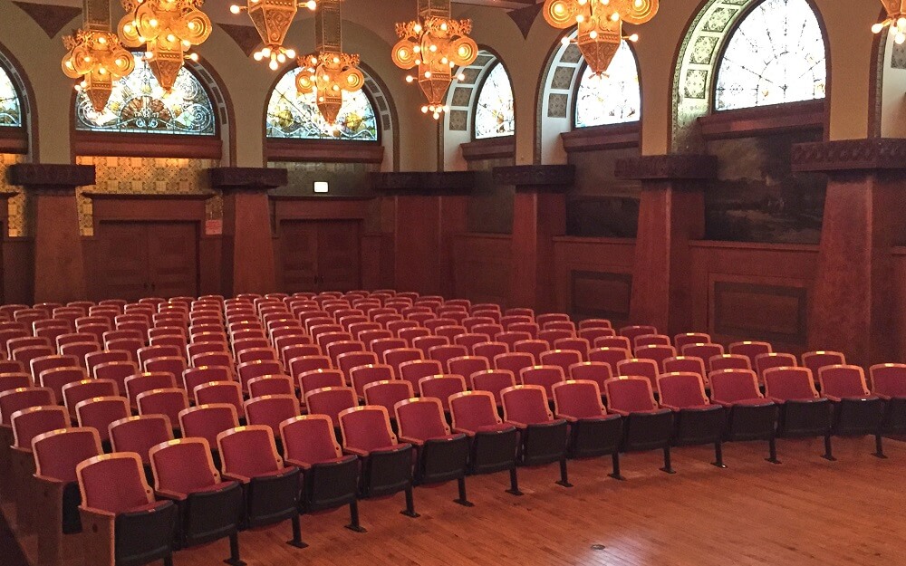 Large room with theater style seating with ornate lighting and stained glass windows.