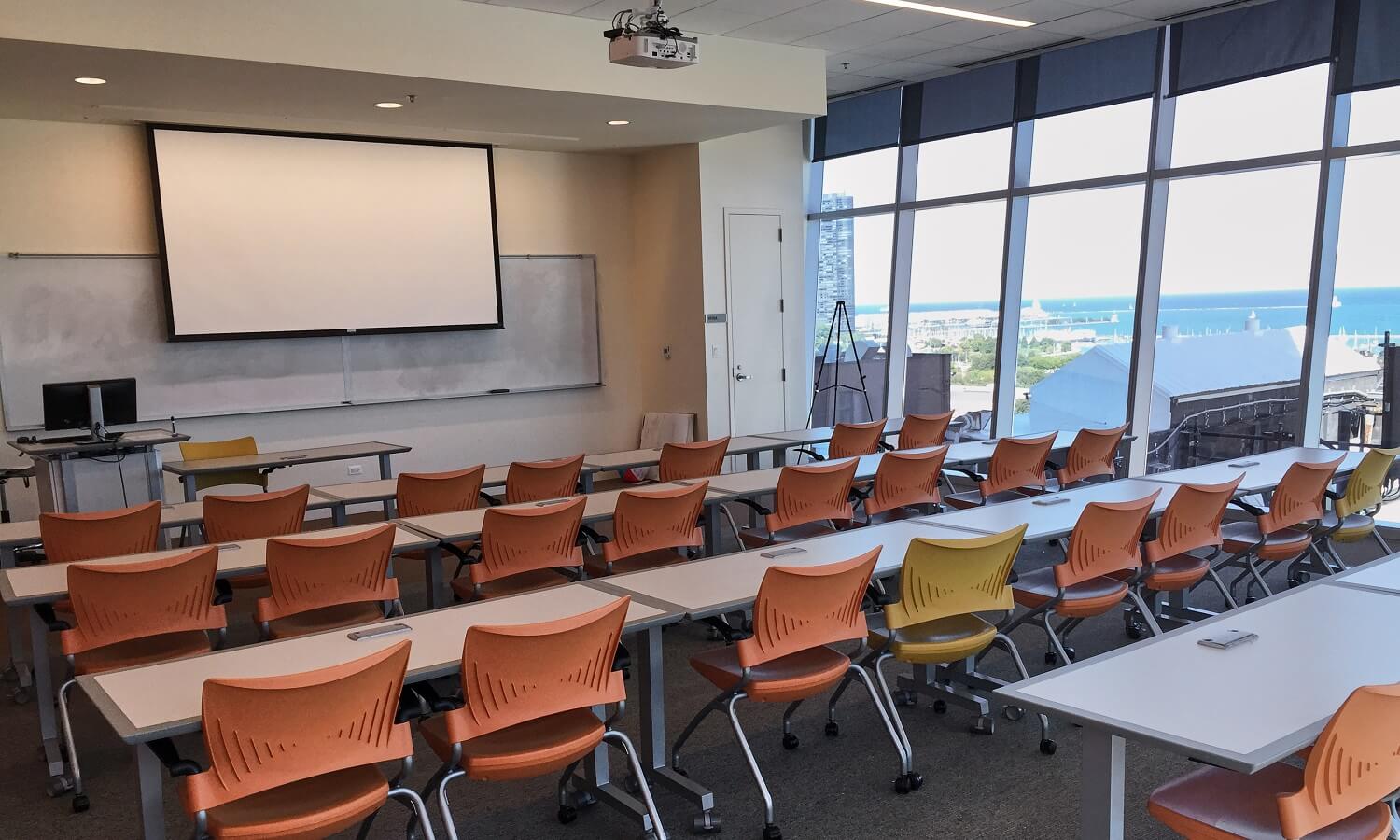 Tiered stadium style room with tables and chairs, white board and floor to ceiling windows.