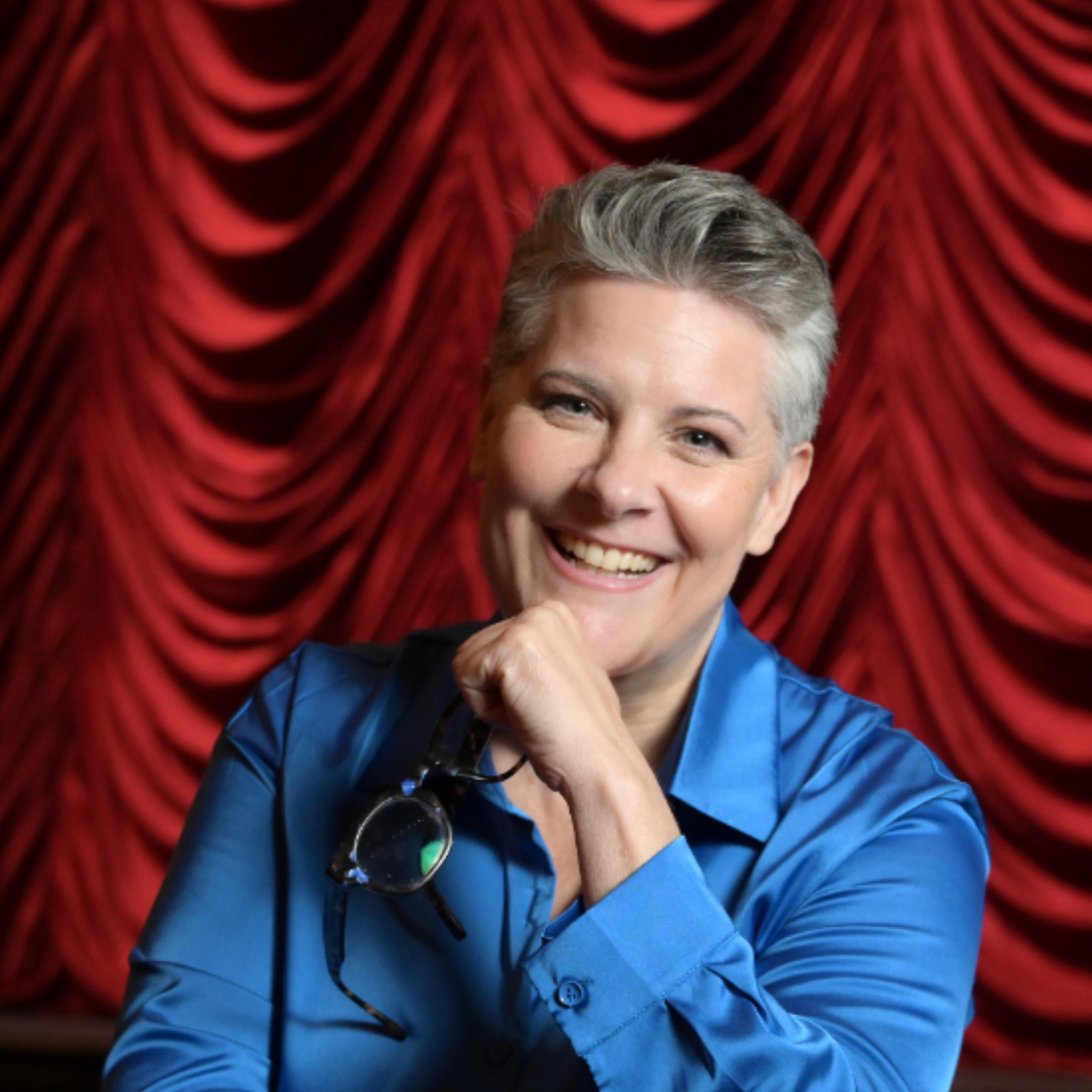 woman stage director smiling in blue blouse in front of red venetian theatrical curtain