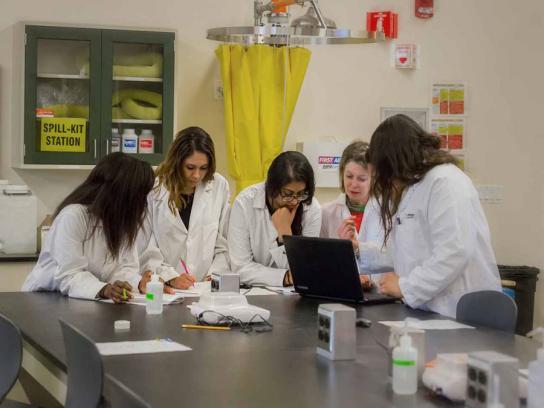 Students in lab coats working together with a laptop and lab equipment.