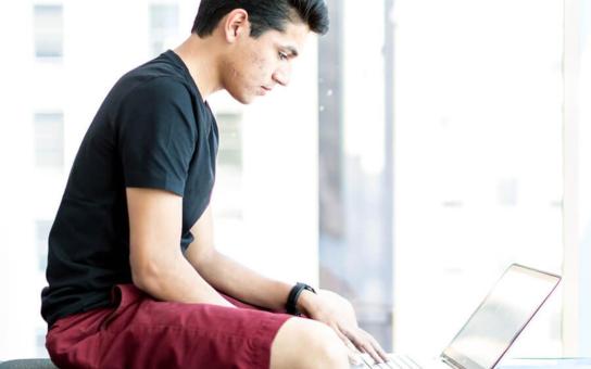 Male student studying by campus windows with a laptop