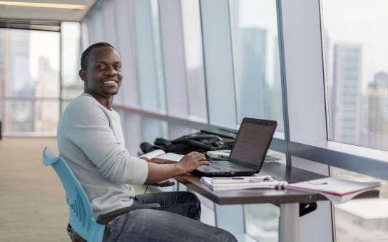 Male student sitting at a desk with an open laptop in a room with a city view