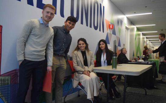 In the foreground, two students standing and two students sitting at table, in the background a student shaking hands with potential employer