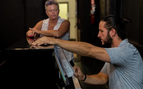 Student sitting at a piano while an instructor stands next to the piano looking at the student