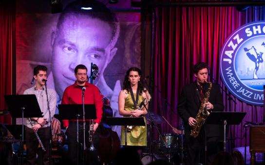 Four students staging on stage holding instruments while one is playing the saxophone.