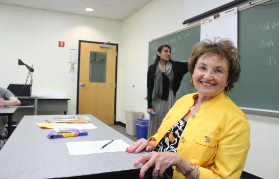 Two women in a classroom, one teaching at a chalkboard the other behind a desk.