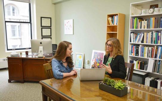 A staff member meeting with a student in a office setting, gesturing with her hand in front of a laptop.