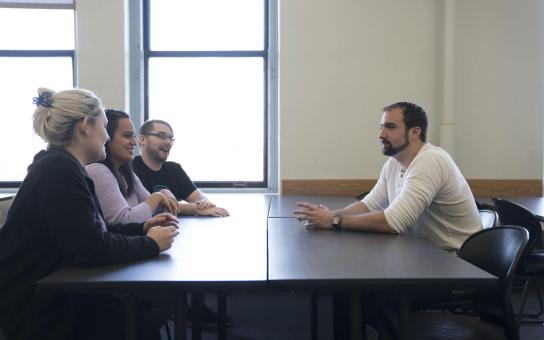 Group of people with three on one side and one on the other in discussion around a square table