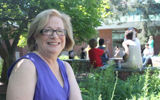 Woman sitting outside smiling an elementary school with students sitting in the far background.