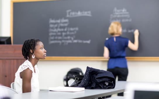 Profile of student sitting at a desk while an instructor writes on a blackboard.