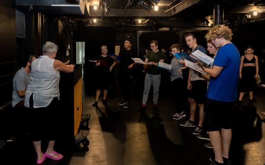 Students standing and singing around a piano in a performance practice room.