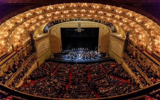 An orchestra performs before an audience on the stage of the Auditorium Theatre