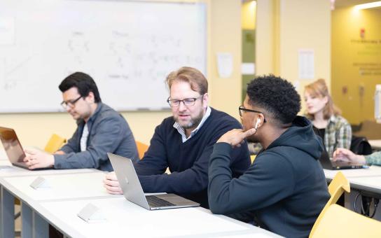 Two individuals work at a laptop in a classroom