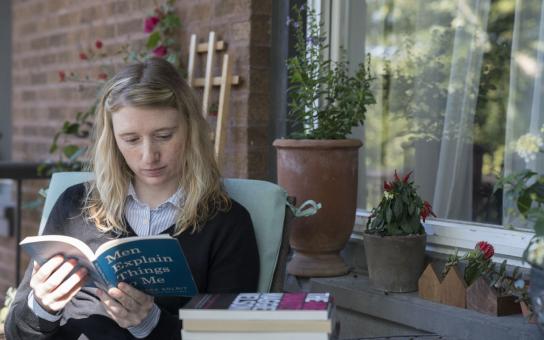 Woman seated outdoors reading a book in front of a window with potted plants on the window sill.