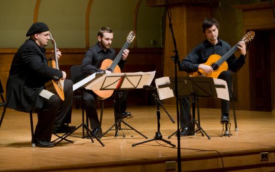 Three students dressed in black, sitting on a stage performing with guitars