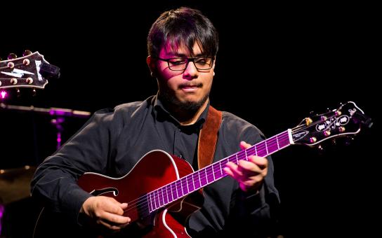 Student in black shirt on a performance stage playing a jazz guitar