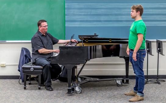 A student standing practicing vocal warm-ups while instructor sits at the piano.