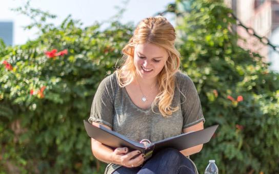 Seated female student reading notes from a binder outdoors in front of landscape