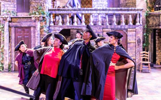 Small group of actors performing on bunched together on stage wearing Victorian era costumes, with hats and capes.