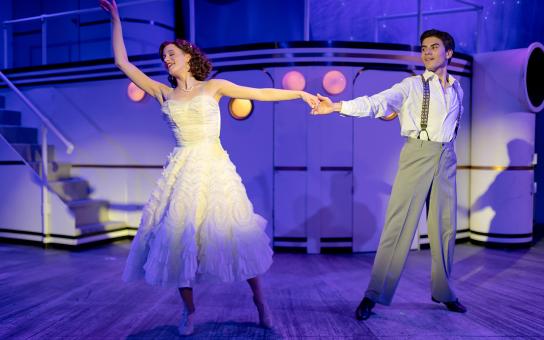 A performer in a white dress and performer in a dress pants hold each other's outstretched arms while dancing on stage