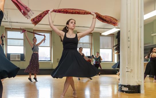 Dancer in black dress holding a scarf over head, while other dancers in the background are doing the same.