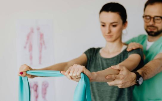 Person with arms extended holding an exercise band while another person is supporting extended arms from behind.