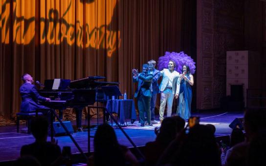 Actors on stage in a spotlight facing a pianist on stage with word Anniversary displayed on the curtain backdrop