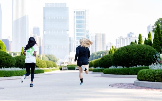 Two students jogging on a paved pathway surrounded by greenery with Chicago cityscape in the background