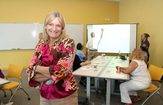 Small group of faculty having a training session in the background while a faculty with arms crossed in smiling at camera in foreground.
