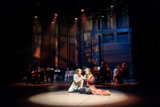 Long shot of two costumed performers kneel facing each other on a spotlighted stage, with musicians in the dimly lit background