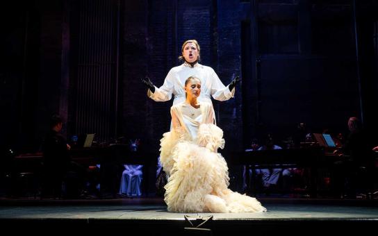 Two performers on stage, one kneeling in a feathered white costume, the other standing with arms raised in white and black.