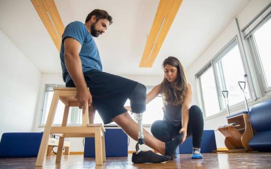 Man sitting in a chair with one leg outstretched and the other prosthetic leg bent while a women kneeling beside him is helping with physical therapy