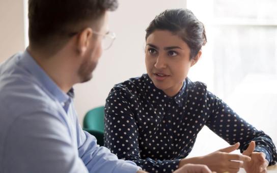 Two people engaged in conversation with one person sitting with hand together actively listening.