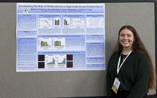Student wearing a "student competition participant" event badge while standing next to a research poster with the title "Investigating The Role of Thiabendazole in High Grade Serous Ovarian Cancer".