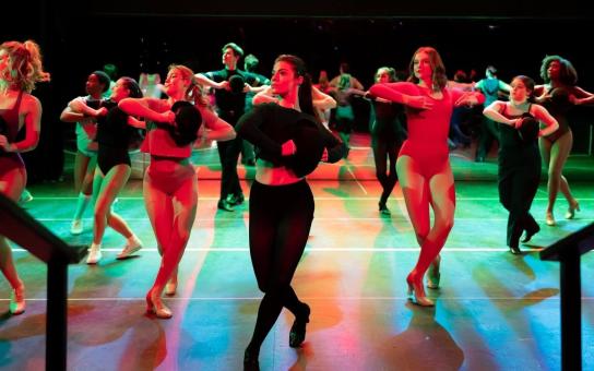 A group of dancers in A Chorus Line rehearses on a stage under colorful red and green lighting, performing a synchronized routine with hats held to their chests. They are arranged in several rows, striking the same pose with strong, coordinated posture.