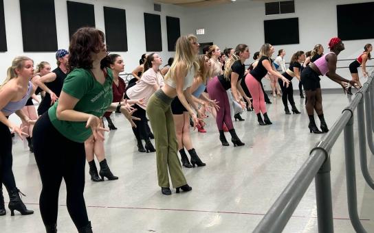 A large group of dancers in a studio practices a synchronized routine for MTD class, bending forward with their arms extended and fingers splayed. Most are wearing dance heels and fitted rehearsal attire, spaced in rows that angle toward a mirrored wall and ballet barres.