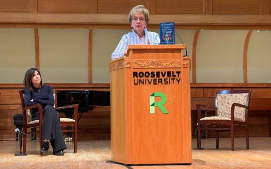 Speaker addresses the audience from a podium as another person sits on stage listening