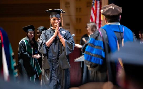 Student in graduation regalia smiles and clasps their hands before accepting their diploma